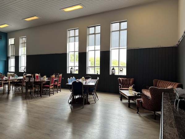 A spacious room with large windows, wooden flooring, and multiple seating areas including tables and chairs.