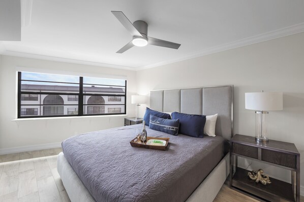 A modern bedroom with a large bed, a ceiling fan, a nightstand with a lamp, and a window with a view of buildings. - Fifth Floor Condo | Pools, Spas, Tennis & Dining (Marco Island)