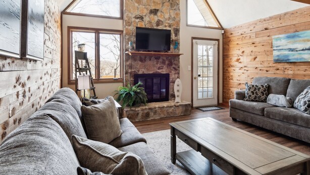 A spacious living room with a stone fireplace, wooden walls, and large windows.