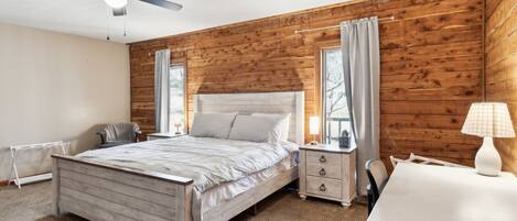 A bedroom with a wooden wall, a bed, a nightstand, a chair, and a desk with a lamp.