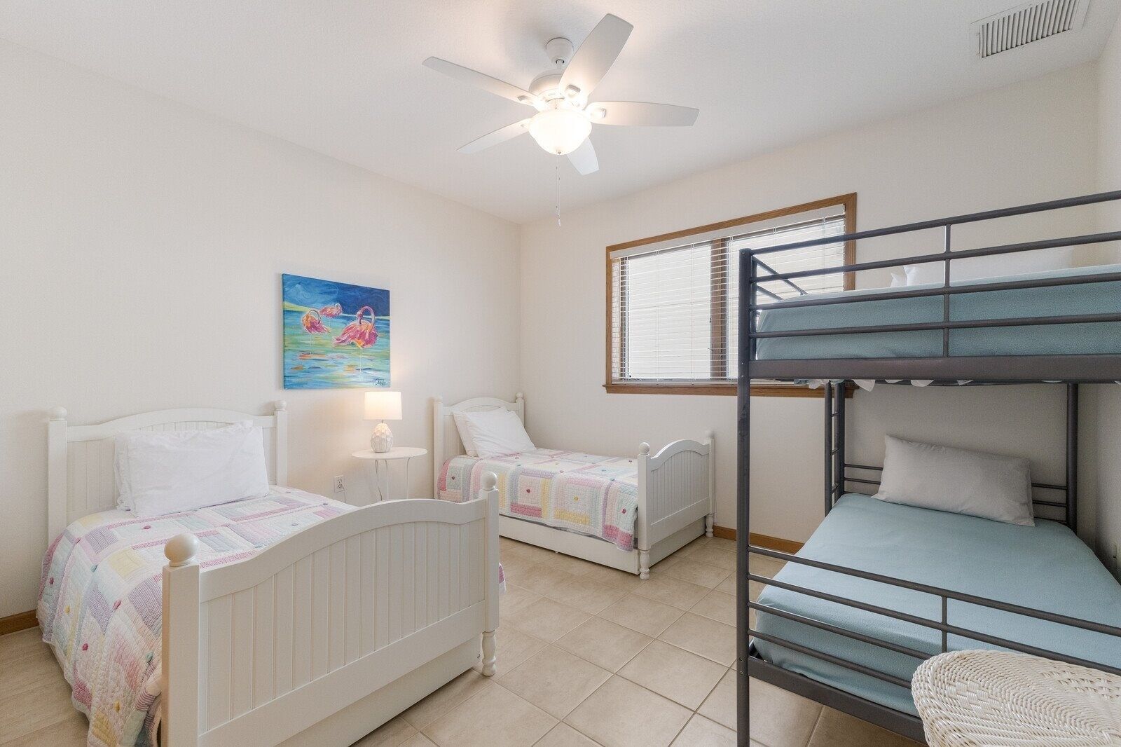 A room with bunk beds, two single beds, a ceiling fan, a window with blinds, and a painting on the wall.