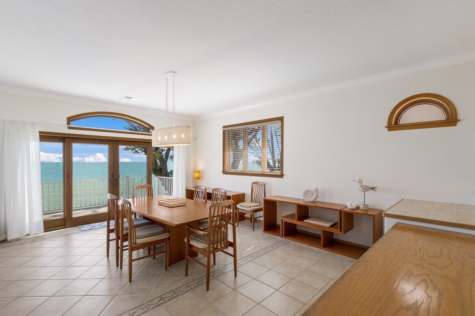 A spacious dining area with a large table and chairs, a hanging light fixture, and a view of the ocean through a sliding glass door.