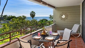 A balcony with a table set for two, overlooking a coastal view with palm trees and the ocean.