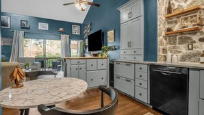 Une cuisine moderne avec des meubles gris, un mur d’accent en pierre et un coin repas comprenant une table en marbre et des chaises noires.
