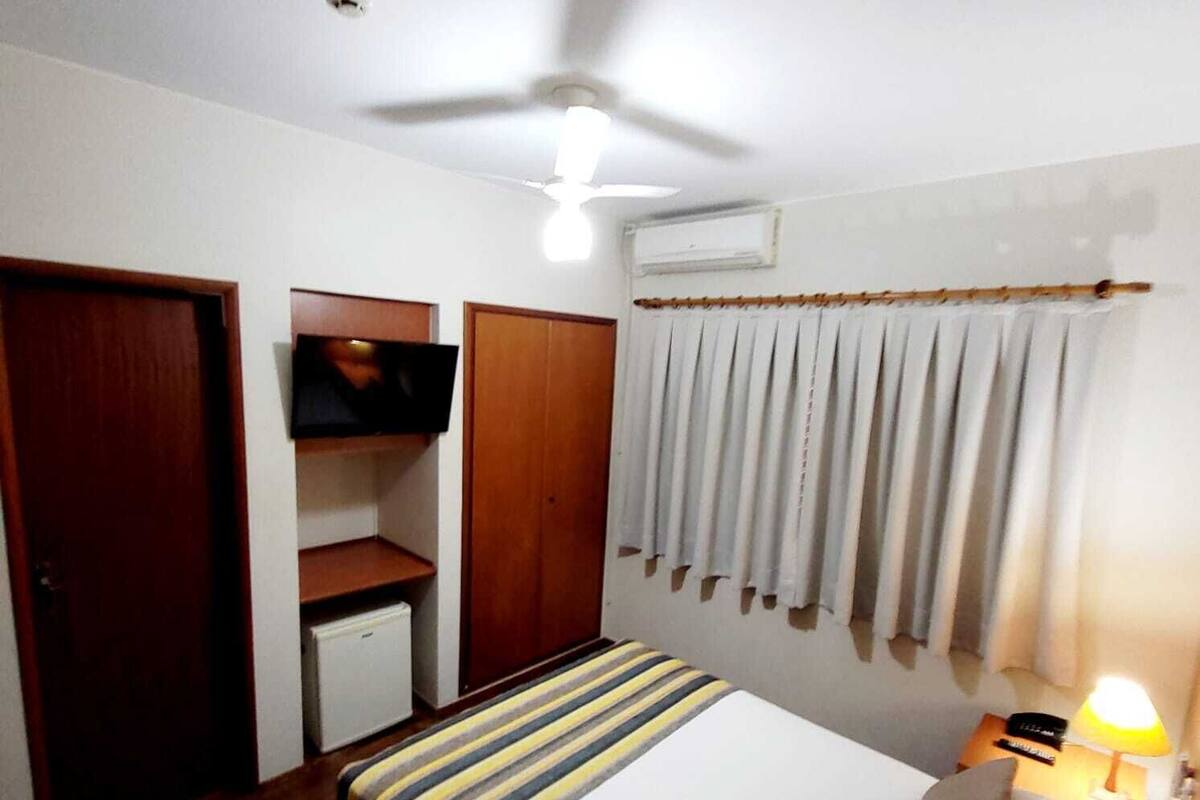 Une chambre à coucher avec un lit, un téléviseur fixé au mur, un ventilateur de plafond et une fenêtre avec des rideaux.