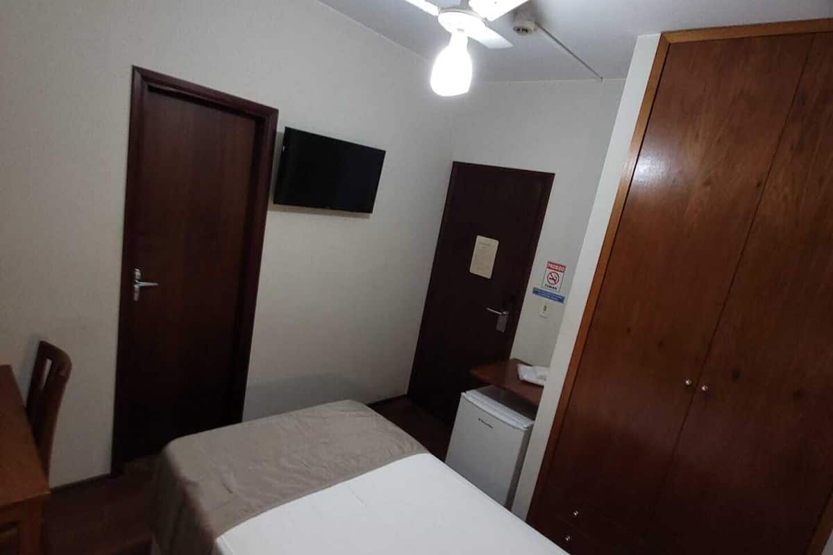 Une petite pièce simple, avec deux lits simples, un bureau, une chaise, une armoire et un ventilateur de plafond.