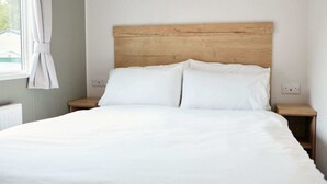 Un lit bien fait, avec du linge de lit blanc, dans une chambre dotée d’une tête de lit en bois, de deux tables de chevet et d’une fenêtre avec des rideaux.