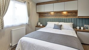 Een netjes opgemaakt bed met een grijze deken, een houten hoofdbord en een nachtkastje met een klok en een fotolijst.