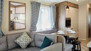 Een moderne woonkamer met een grijze hoekbank, een eethoek met een houten tafel en stoelen, en een keuken die zichtbaar is door een glazen wand.