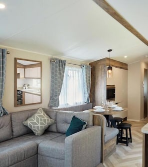 Ein modernes Wohnzimmer mit einem grauen Ecksofa, einem Essbereich mit Holztisch und -stühlen sowie einer durch eine Glaswand sichtbaren Küche.