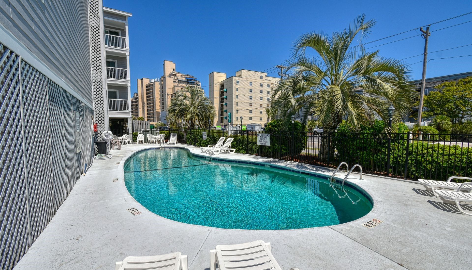 A swimming pool with lounge chairs and a fence, surrounded by buildings and palm trees.