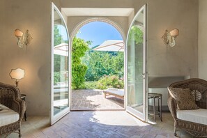 An open patio with a glass door leading to a lush garden, wicker furniture, and a white umbrella. - Col Di Casale - A breathtaking Umbrian villa in the green heart of Italy. (Pietralunga)