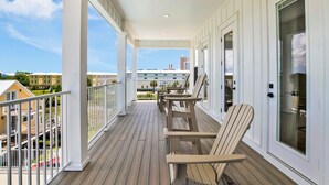 Un balcón techado al aire libre con piso de madera, barandillas blancas y dos sillas Adirondack.