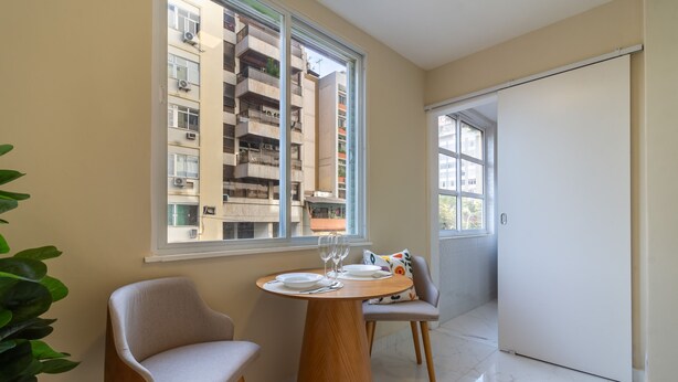 A modern dining area with a round table, two chairs, and a view of a high-rise building through a large window. - Comfort in Ipanema 6 Minutes From the Beach J302 (Rio de Janeiro)