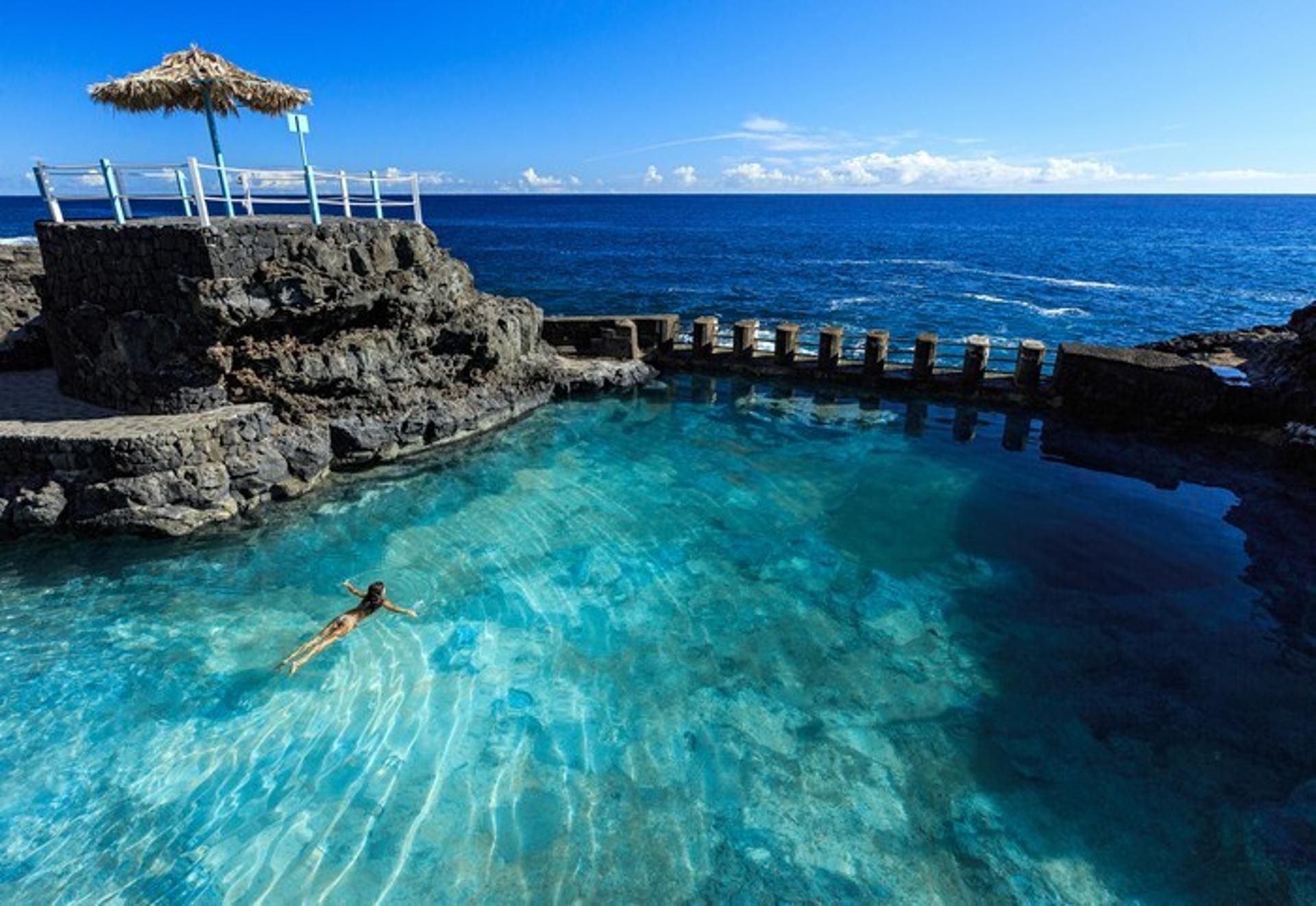 En klarblå pool med en person som simmar i den, omgiven av ett bergigt landskap och med utsikt över havet.