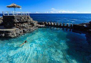 Une piscine d’un bleu éclatant où une personne nage, entourée d’un paysage rocheux et offrant une vue sur l’océan.