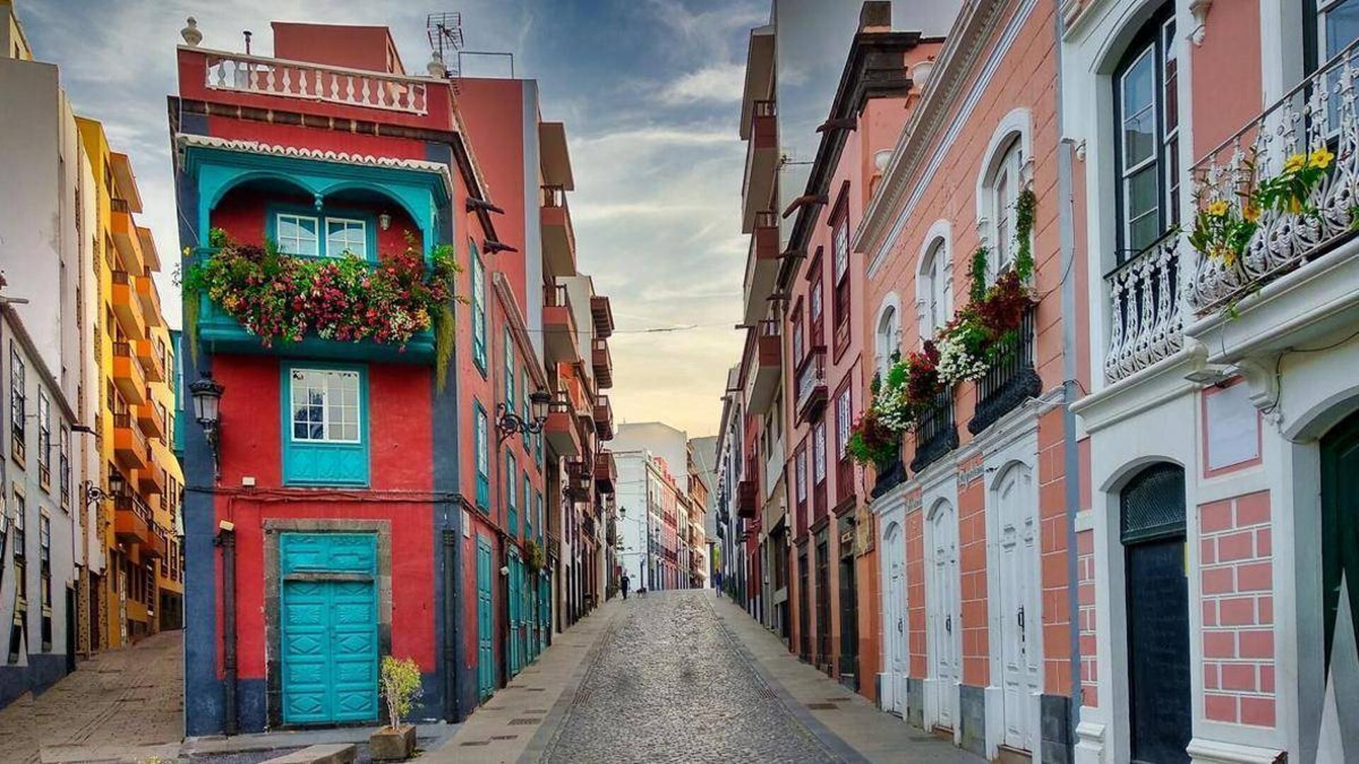 A narrow cobblestone street lined with colorful buildings, some with balconies and flower boxes.