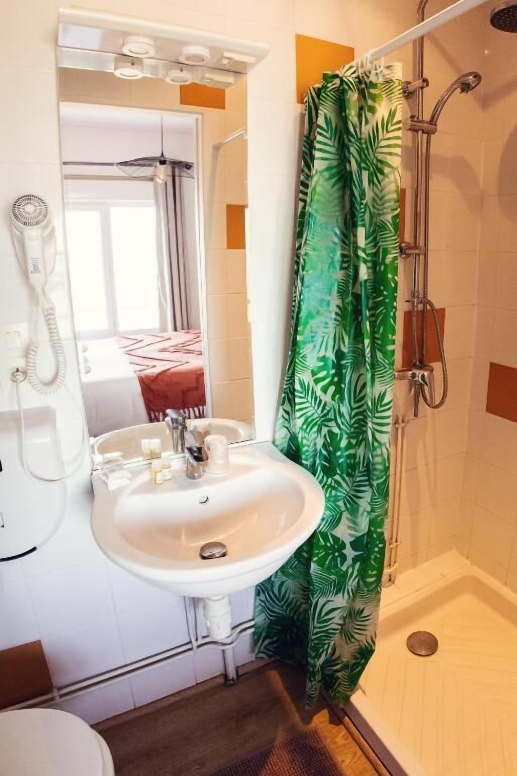 Un baño con un lavabo blanco, un espejo, una ducha con cortina estampada con motivos de hojas verdes y un teléfono en la pared.