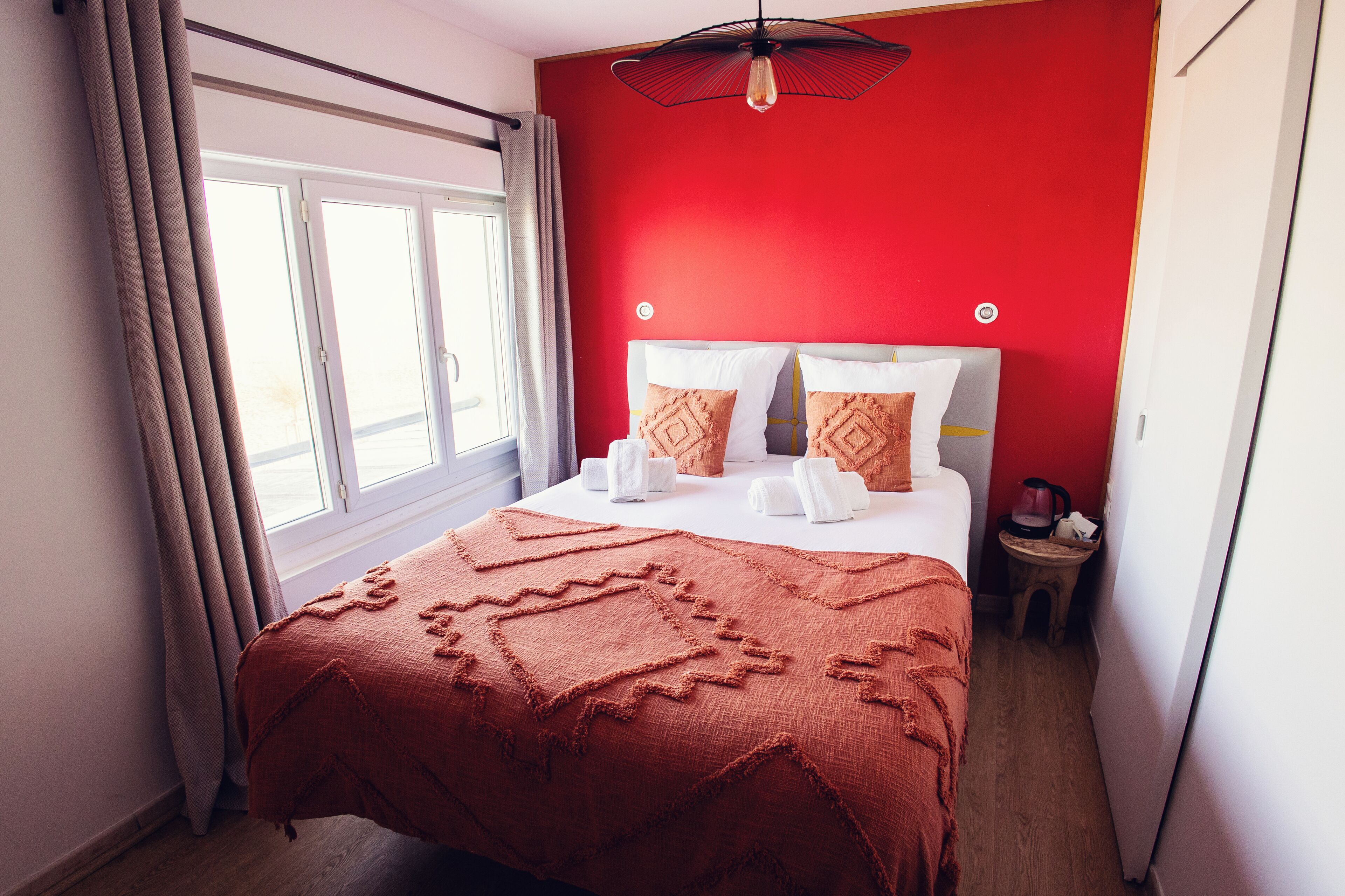 Habitación de hotel con una pared de acento roja, una cama grande con colcha estampada, dos almohadas y una mesita de noche con tetera.