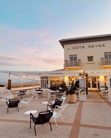 Un área de estar al aire libre con mesas y sillas, un edificio con un letrero que dice "LA COTE REVEE", y una vista de la playa al fondo.