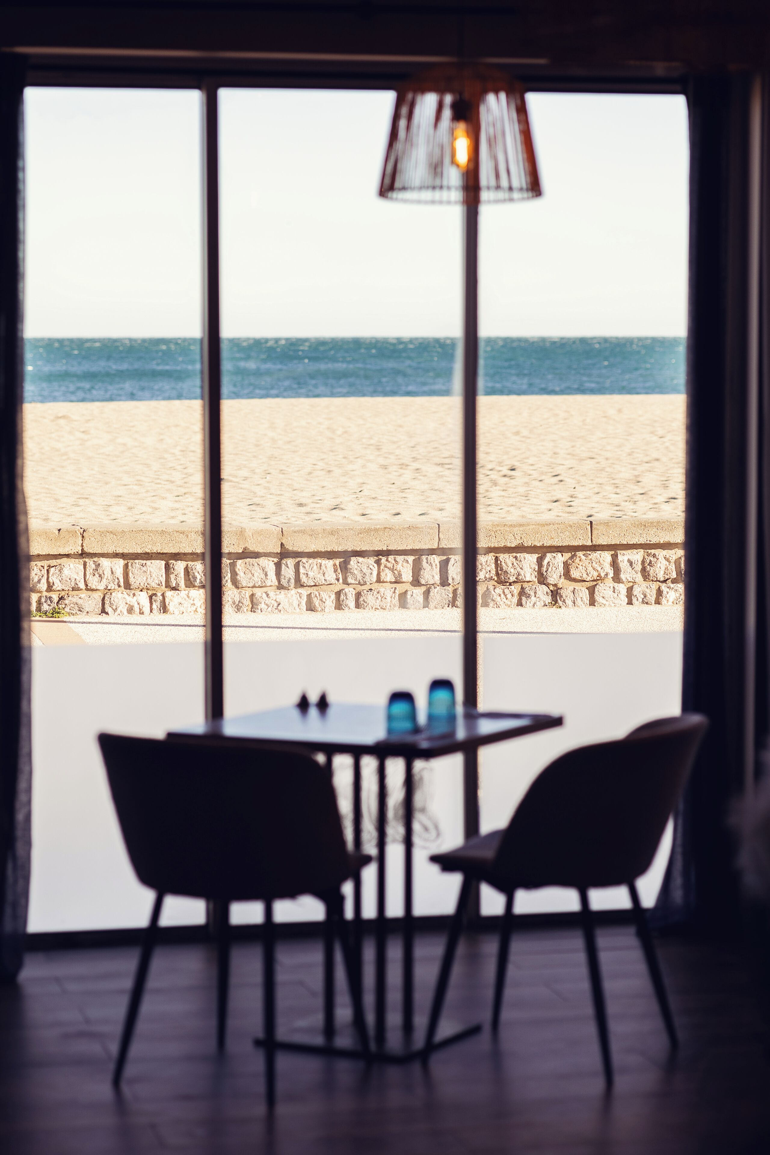 A room with a view of a beach through large windows, featuring a table and chairs, and a hanging lamp.