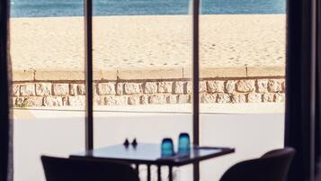 A room with a view of a beach through large windows, featuring a table and chairs, and a hanging lamp.