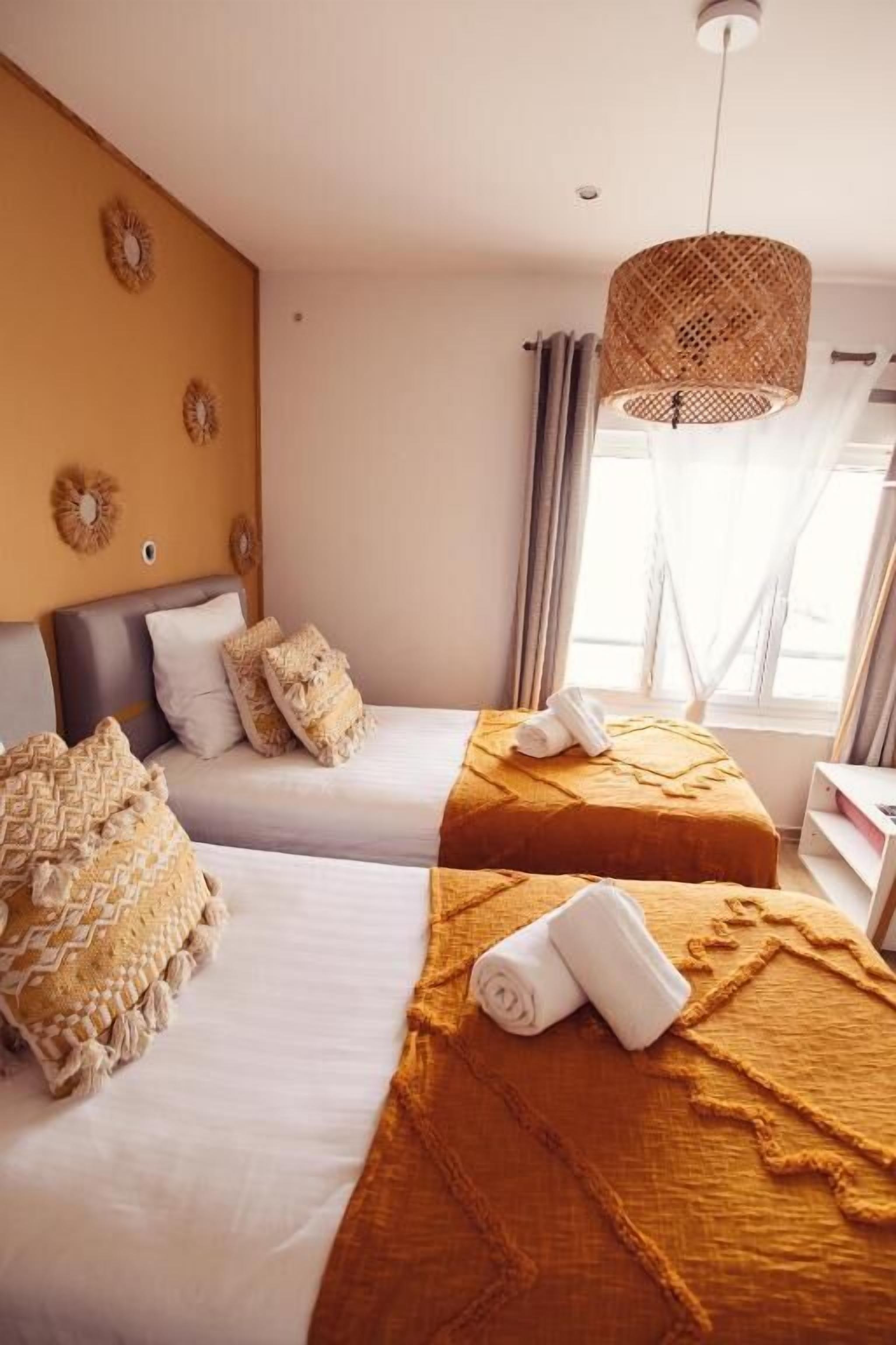 A hotel room with two beds, a large window with curtains, a woven light fixture, and decorative wall mirrors.