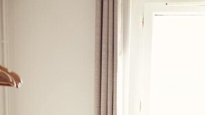 A corner of a room with a white chair, a white wall, and a window with curtains. - La Côte Révéé (Leucate)