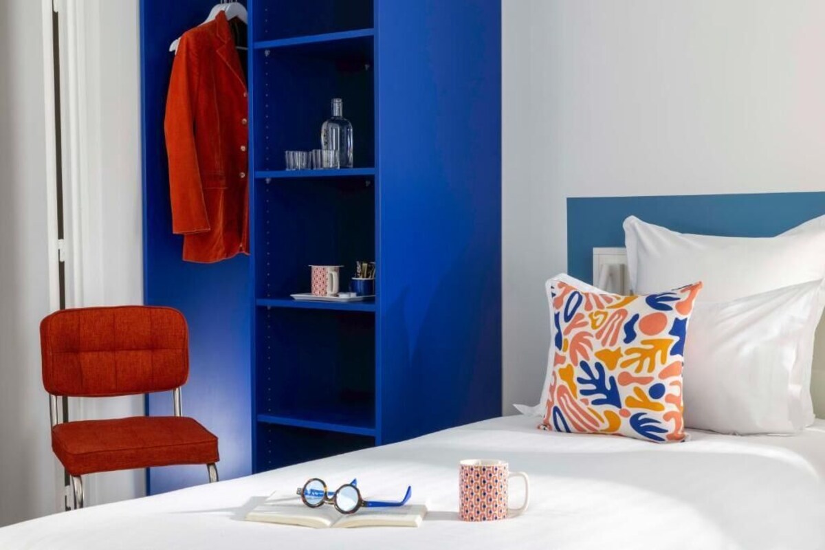 Une pièce avec un lit, un fauteuil rouge, une bibliothèque bleue et un coussin à motifs.