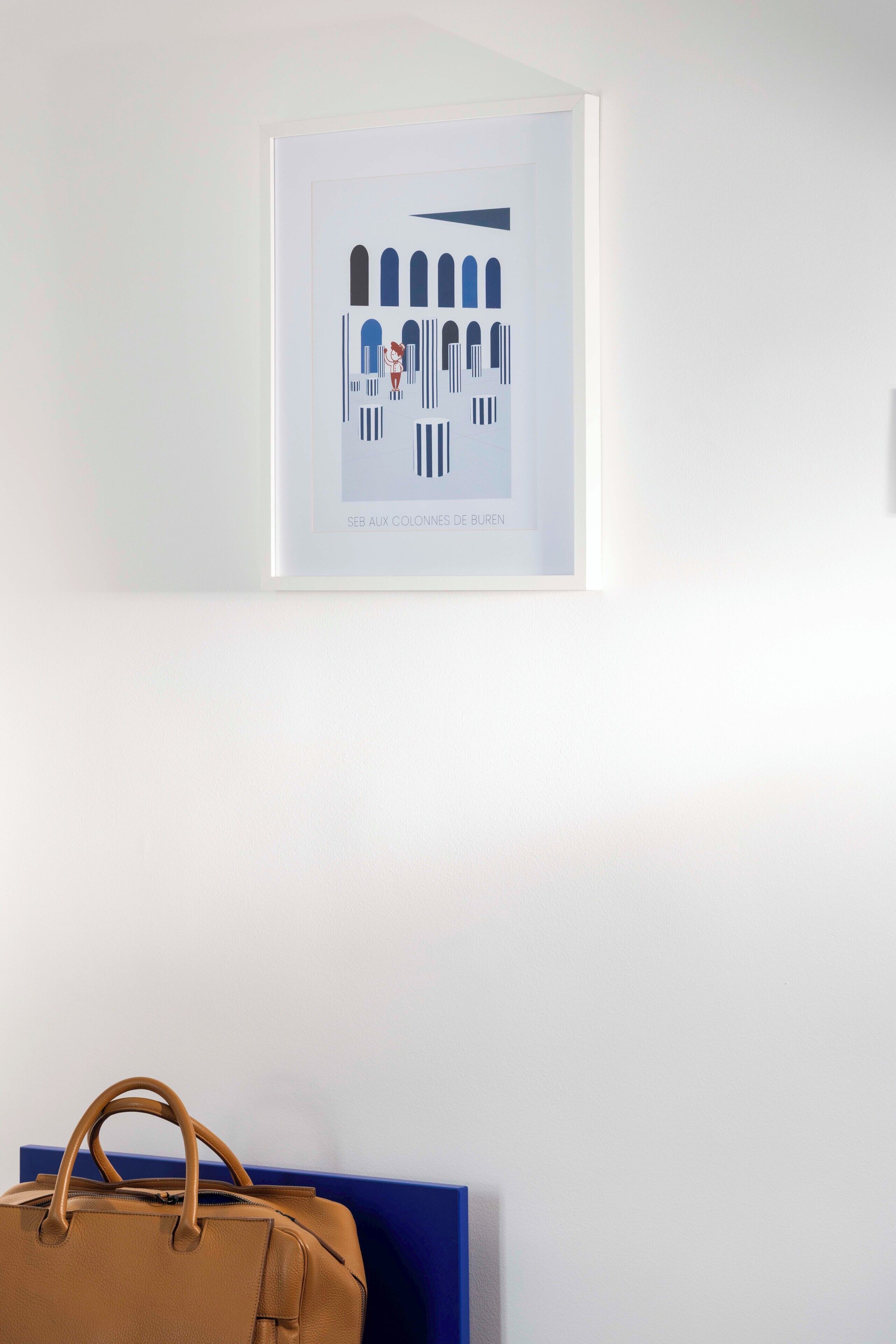 A framed artwork depicting a building with arches and columns, and a brown handbag on a blue bench below.