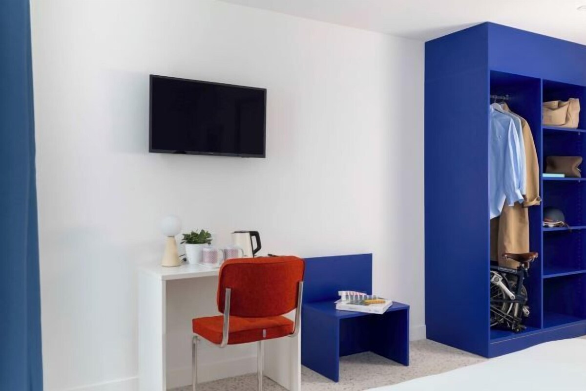 A room with a TV mounted on the wall, a red chair, a small table with a plant, and a blue shelving unit with clothes.