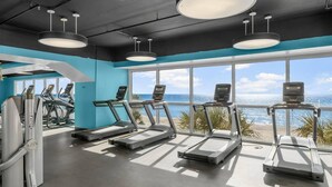 Un gimnasio con cintas de correr y equipo de ejercicio, amplias ventanas con vistas a la playa.