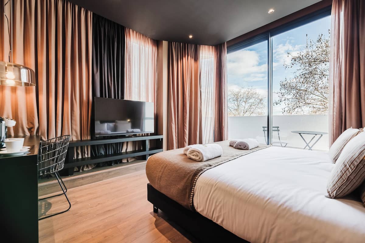 A modern hotel room with a large bed, a flat-screen TV, a desk with a lamp, and a balcony with a table and chairs.