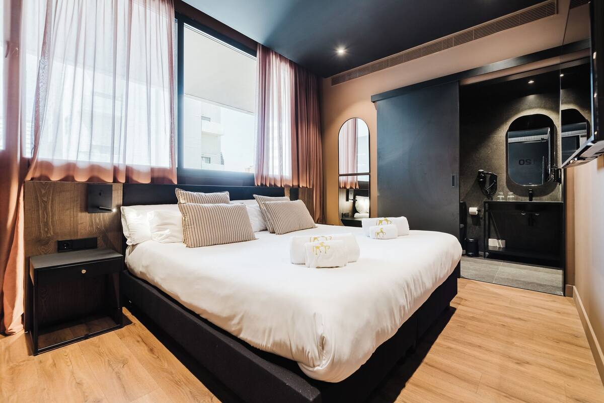 A modern hotel room with a large bed, wooden flooring, a nightstand, a mirror, and a bathroom with a shower and toilet.
