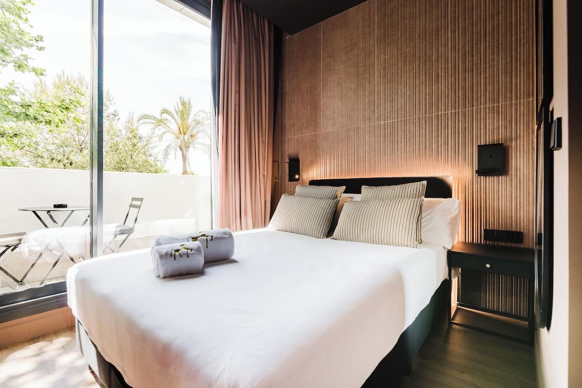 A modern hotel room with a large bed, a nightstand, a wall-mounted TV, and a view of palm trees through the window.