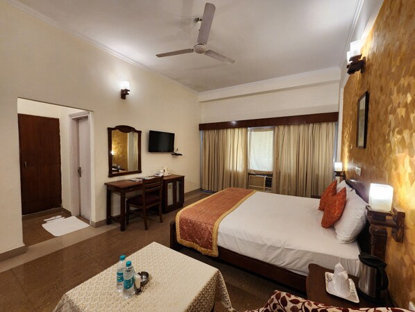 A hotel room with a bed, a desk, a chair, a television, and a ceiling fan. - Sunbird Tourist Resort (Faridabad)