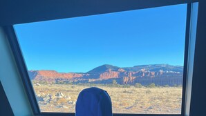 Une personne portant un sweat à capuche bleu se tient dans une pièce éclairée par une lucarne et contemple un paysage de montagnes sous un ciel dégagé.