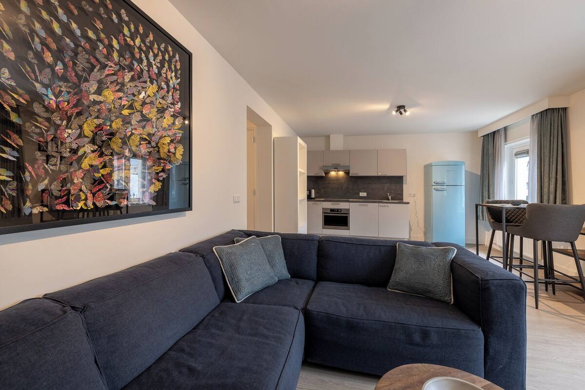 Een moderne woonkamer met een hoekbank, een eethoek met een tafel en stoelen, en een keuken op de achtergrond.