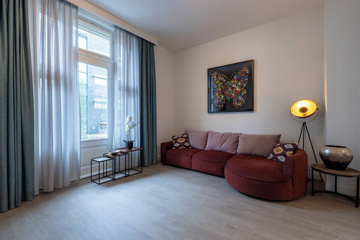 Een moderne woonkamer met een rode bank, een vloerlamp en een groot raam met gordijnen.