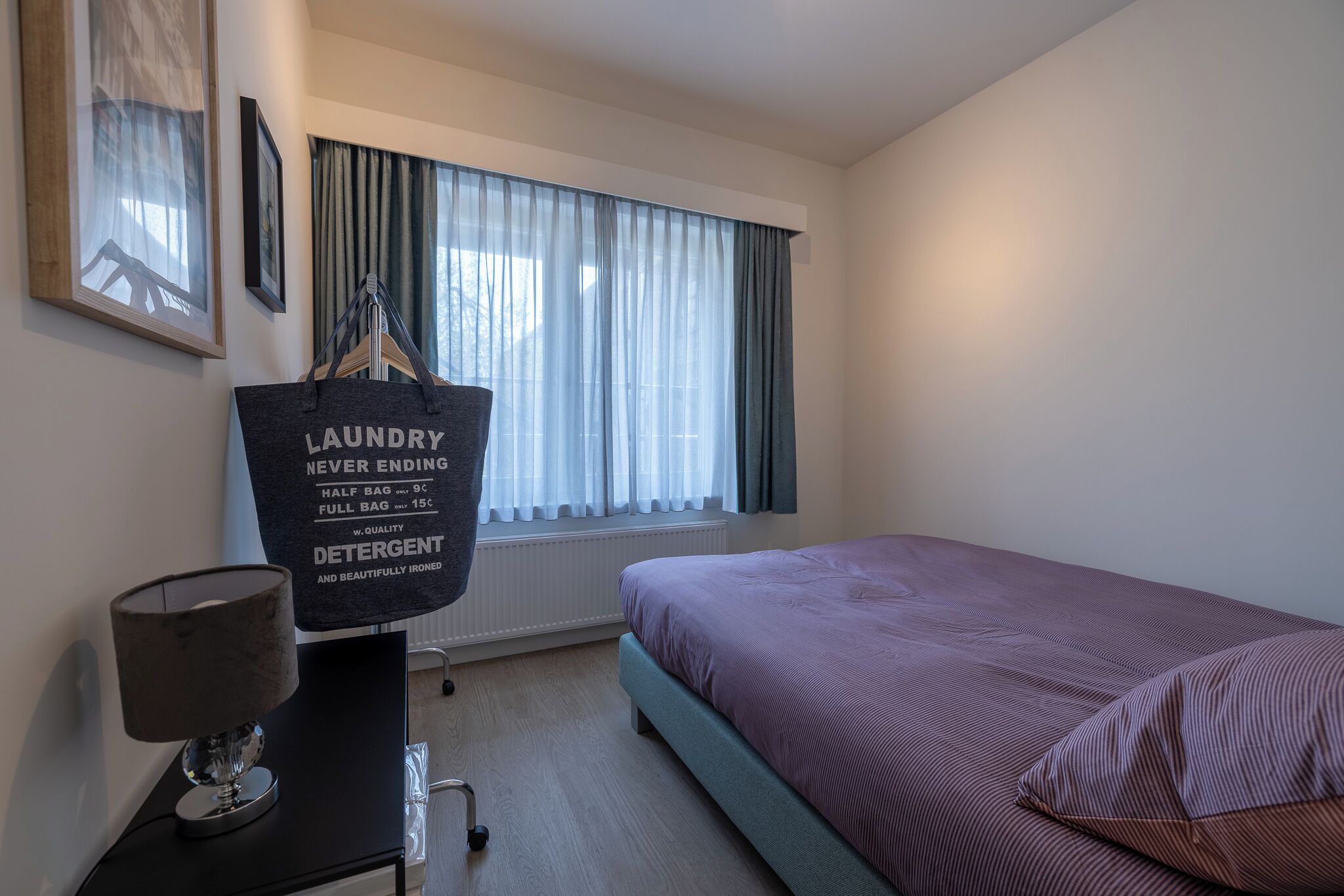 Habitación de hotel con cama, escritorio con lámpara, ventana con cortinas y bolsa para ropa colgada en la pared.