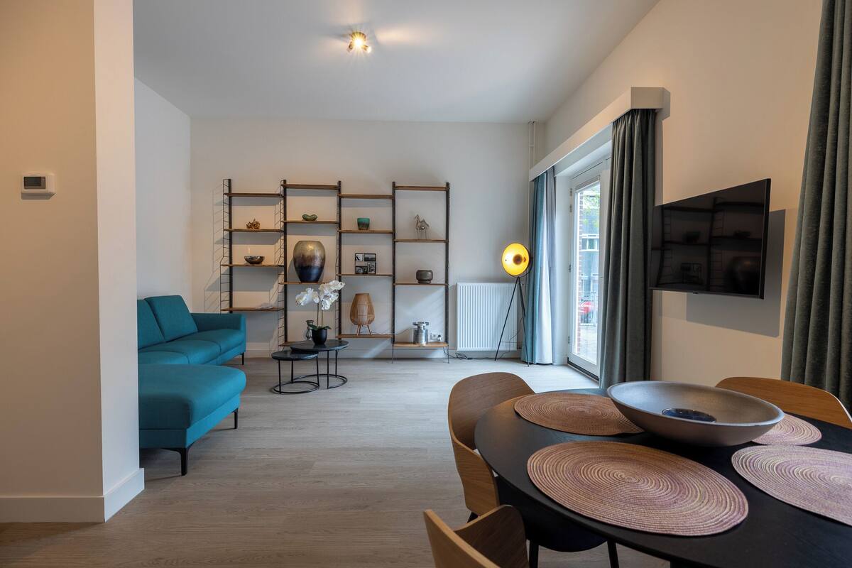 Een moderne woonkamer met een blauwgroene bank, een eettafel met placemats, en een decoratieve plank met diverse items.