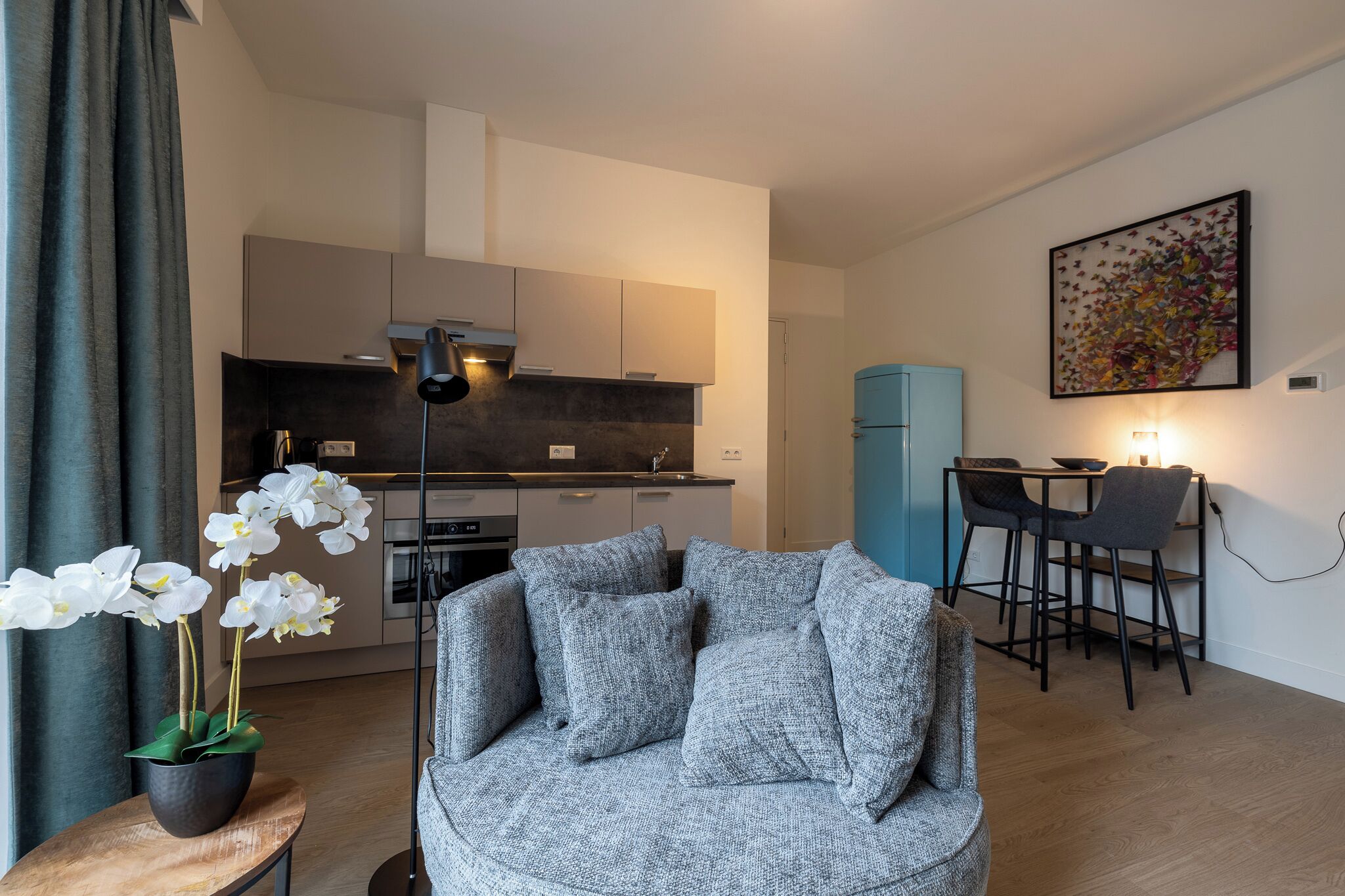 Un salon moderne avec un canapé gris, une petite table sur laquelle est posée une orchidée en pot, et un coin repas comprenant deux chaises et un buffet bleu.