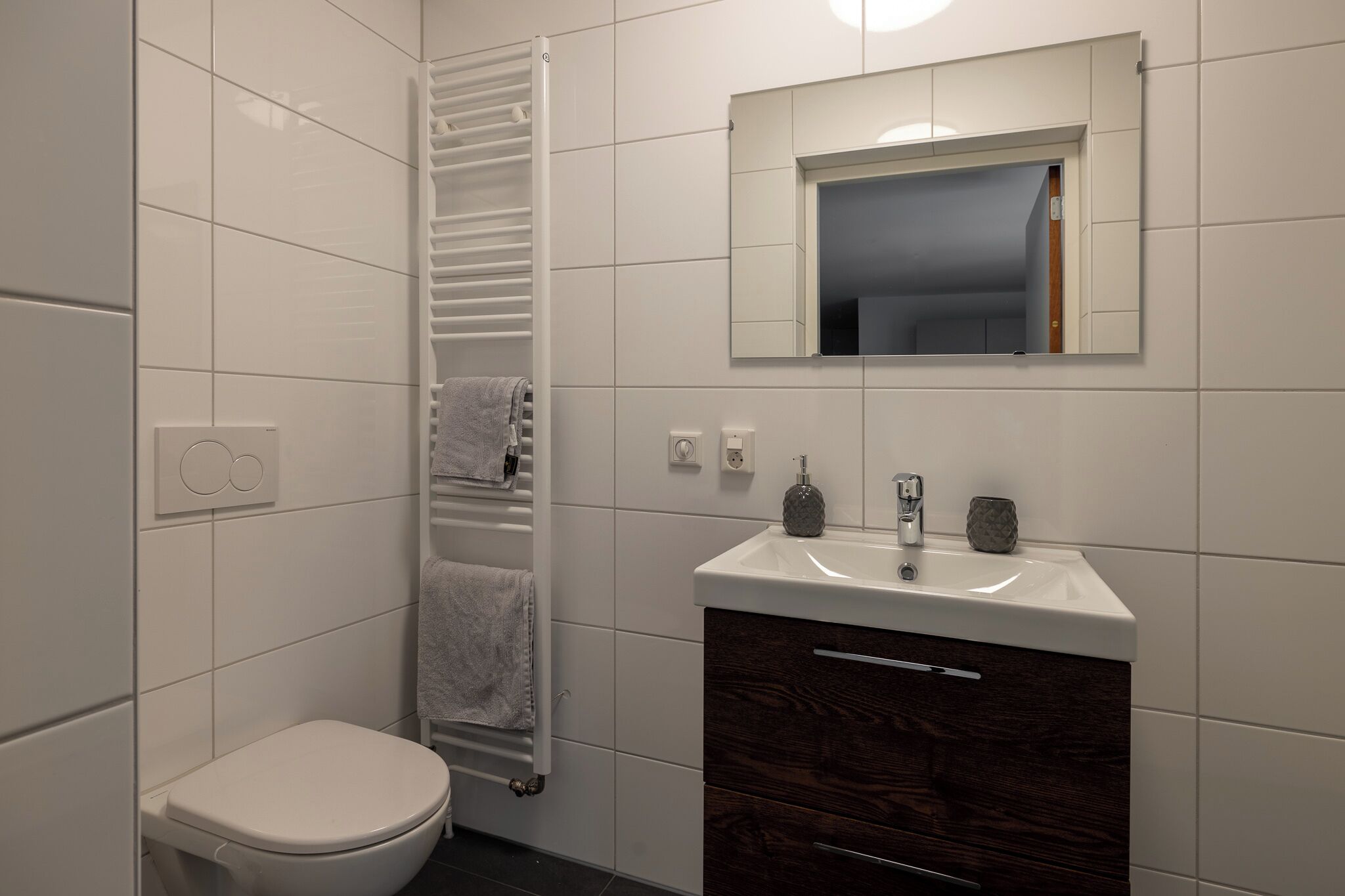 A modern bathroom with white tiles, a white toilet, a towel rack, a sink with a mirror above it, and a dark wood vanity.
