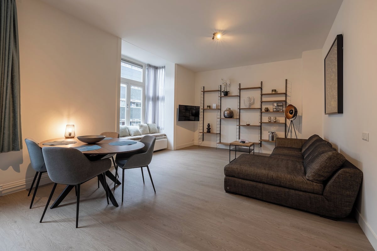 Een moderne woonkamer met een eethoek, een bank en een aan de muur bevestigde televisie.