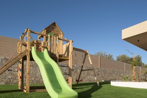 Eine hölzerne Spielkonstruktion mit Rutsche, Kletterwand und Schaukeln vor einer Steinmauer und blauem Himmel.