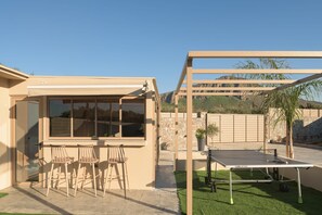 A patio area with a table tennis table, three wooden chairs, and a pergola structure.