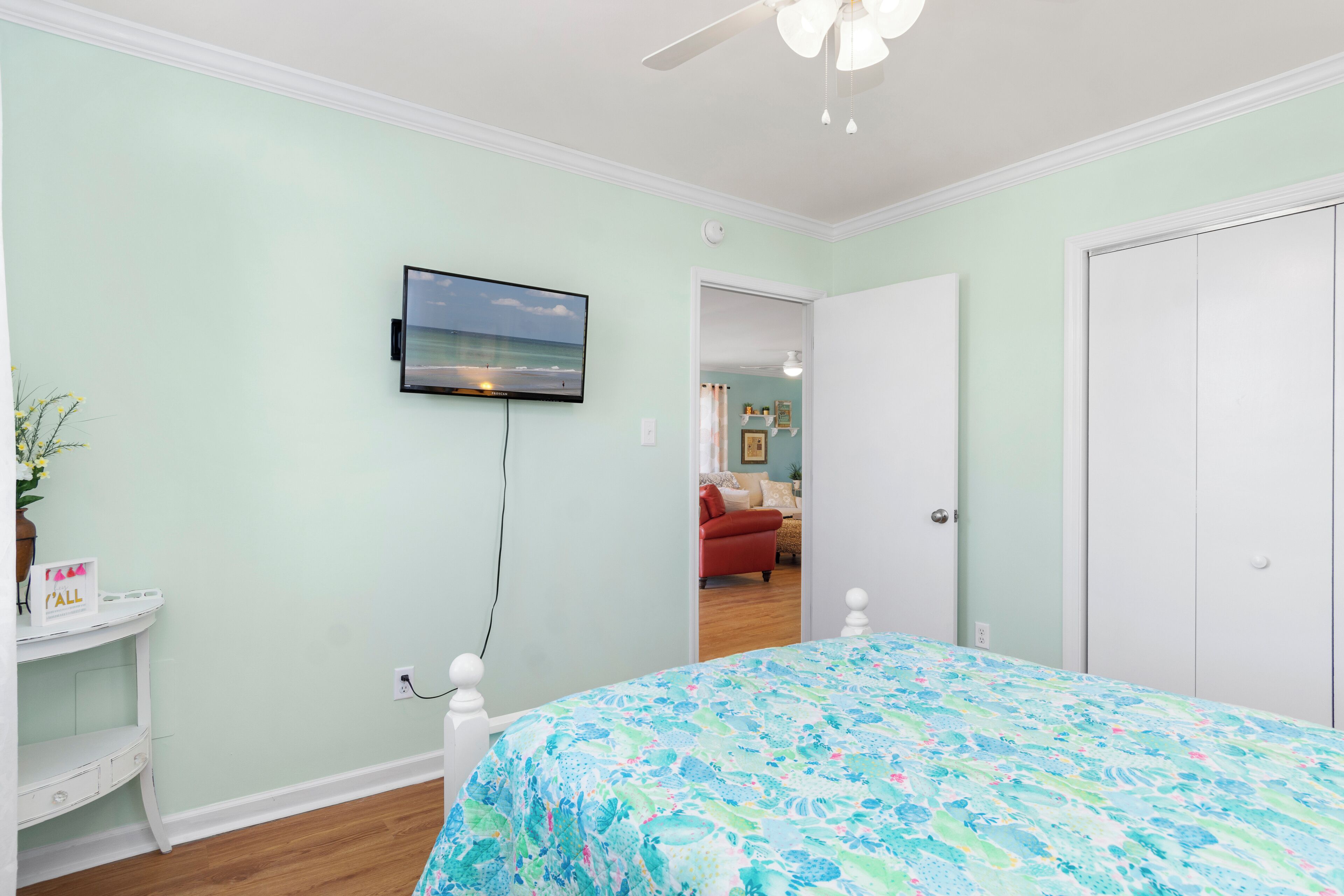 A bedroom with a bed, a TV mounted on the wall, a small table with a vase, and a door leading to another room.