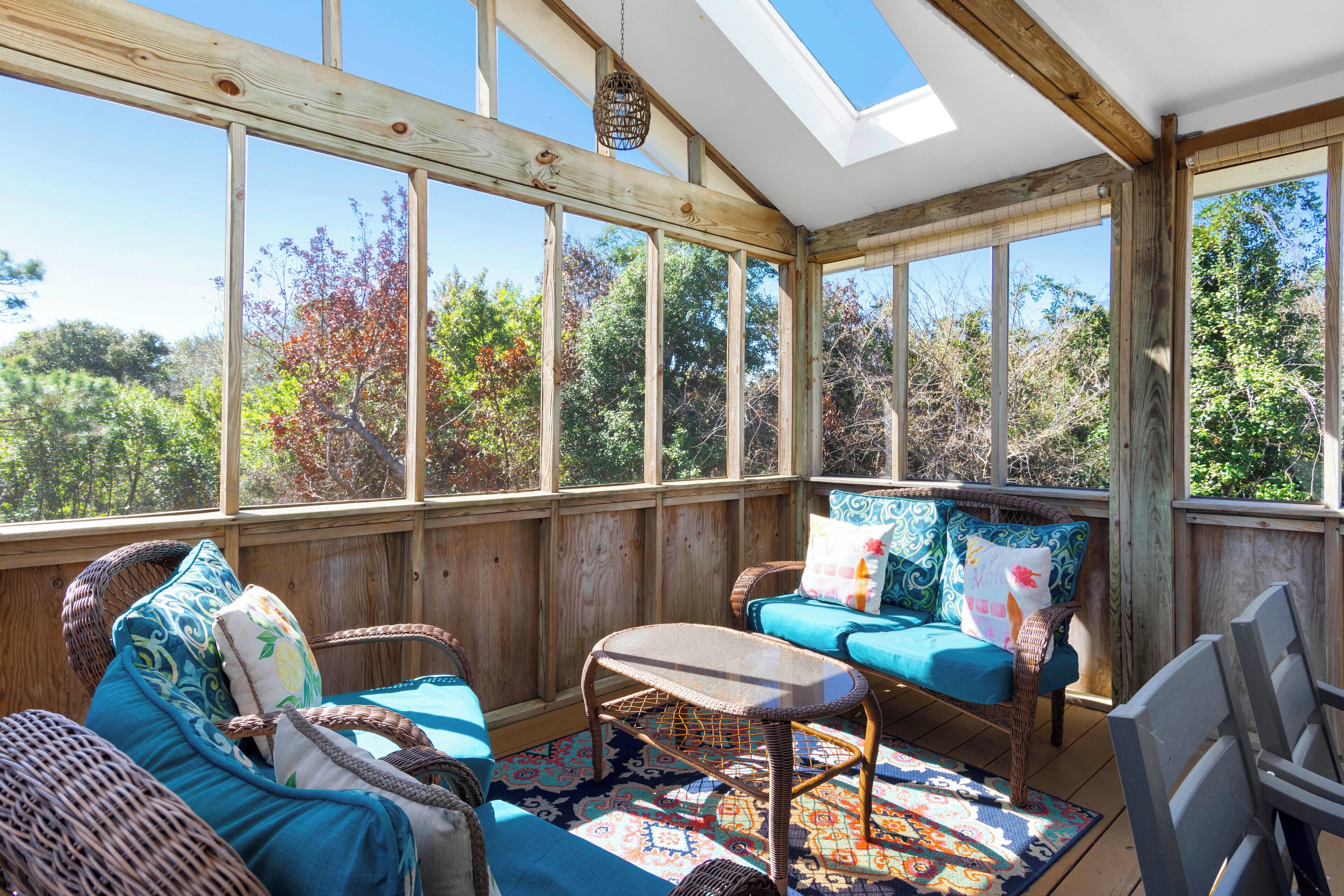 A sunroom with wicker furniture, a patterned rug, and large windows offering a view of trees.