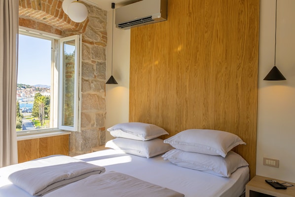 A bedroom with a stone wall, wooden headboard, white bedding, and a view of a town through a window. - East Coast Deluxe Rooms (Split)