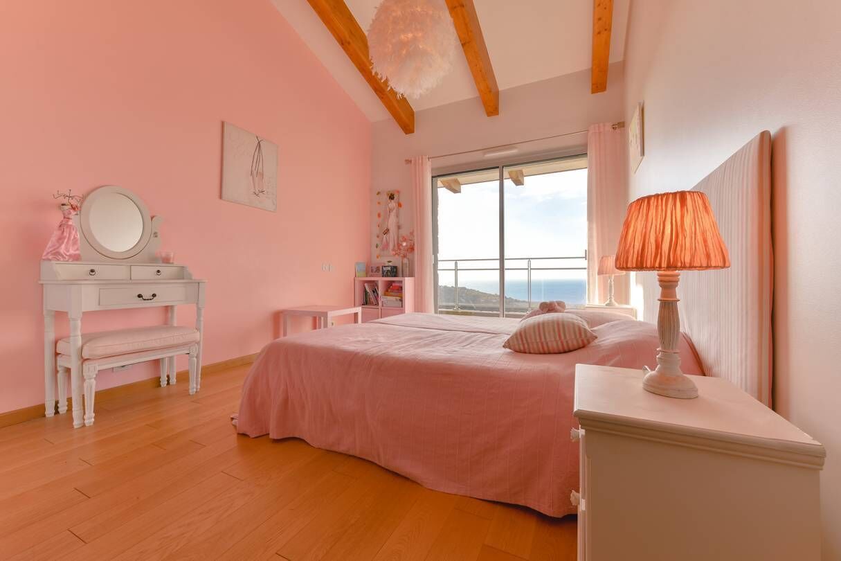 Une chambre à coucher décorée dans des tons roses, avec un lit, une coiffeuse dotée d’un miroir et une fenêtre offrant une vue agréable.